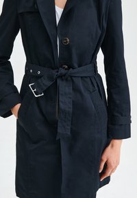 Námořnický trench coat s páskem v pase, s knoflíkovým zapínáním, bočními kapsami a hladkou, jemnou texturou látky.