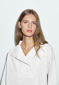 Weißes Oversized-Hemd mit tiefem Kragen, ausgestattet mit einer Knopfleiste und lockerer Passform. Weicher Baumwollstoff mit glatter Textur.