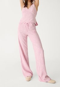 Femme posant dans un haut sans manches rose avec broderies florales et un pantalon large assorti, portant des chaussures à enfiler en toile beige.