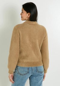 Pull marron en laine duveteuse avec un col montant et des manches ballon. Porté avec un jean en denim bleu légèrement usé. Vue de dos avec une texture lisse.