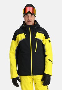 Gelbe und schwarze Skijacke mit einer Reißverschlusstasche, reflektierenden Akzenten und verstellbarer Kapuze, kombiniert mit gelben Hosen und schwarzen Handschuhen.