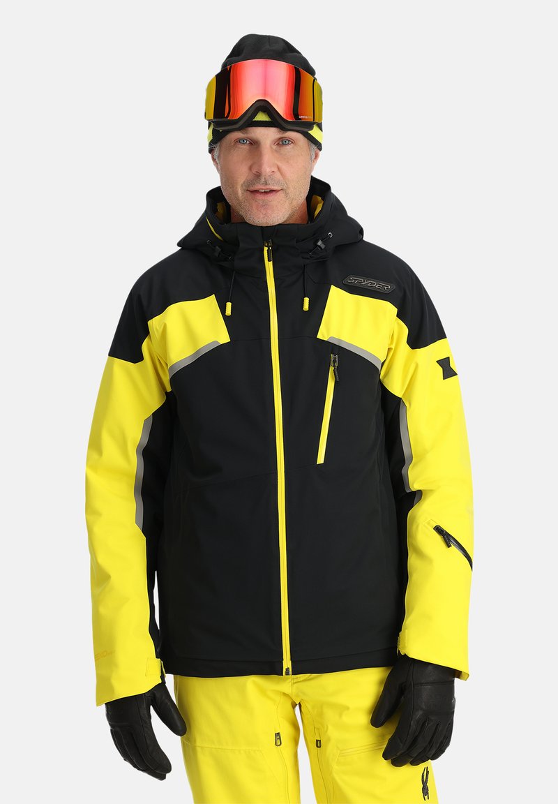 Gelbe und schwarze Skijacke mit einer Reißverschlusstasche, reflektierenden Akzenten und verstellbarer Kapuze, kombiniert mit gelben Hosen und schwarzen Handschuhen.