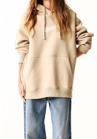Beige Übergröße-Hoodie mit einer verstellbaren Kapuze, Kängurutasche und gerippten Bündchen. Hat ein weißes Logo auf der Brust. Kombiniert mit blauen Jeans.