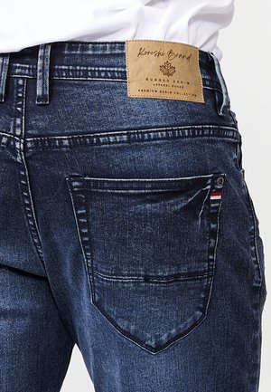 Dunkelblaue Denim-Jeans mit geradem Schnitt, ausgestattet mit einem Leder-Logo-Patch am Bund und rot, weiß und blauem Stich auf der Tasche.