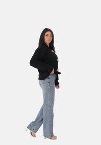 Donna con lunghe capelli scuri che indossa un cardigan nero peloso con grandi bottoni, jeans azzurri chiari e sandali con tacco trasparente mentre cammina di lato.