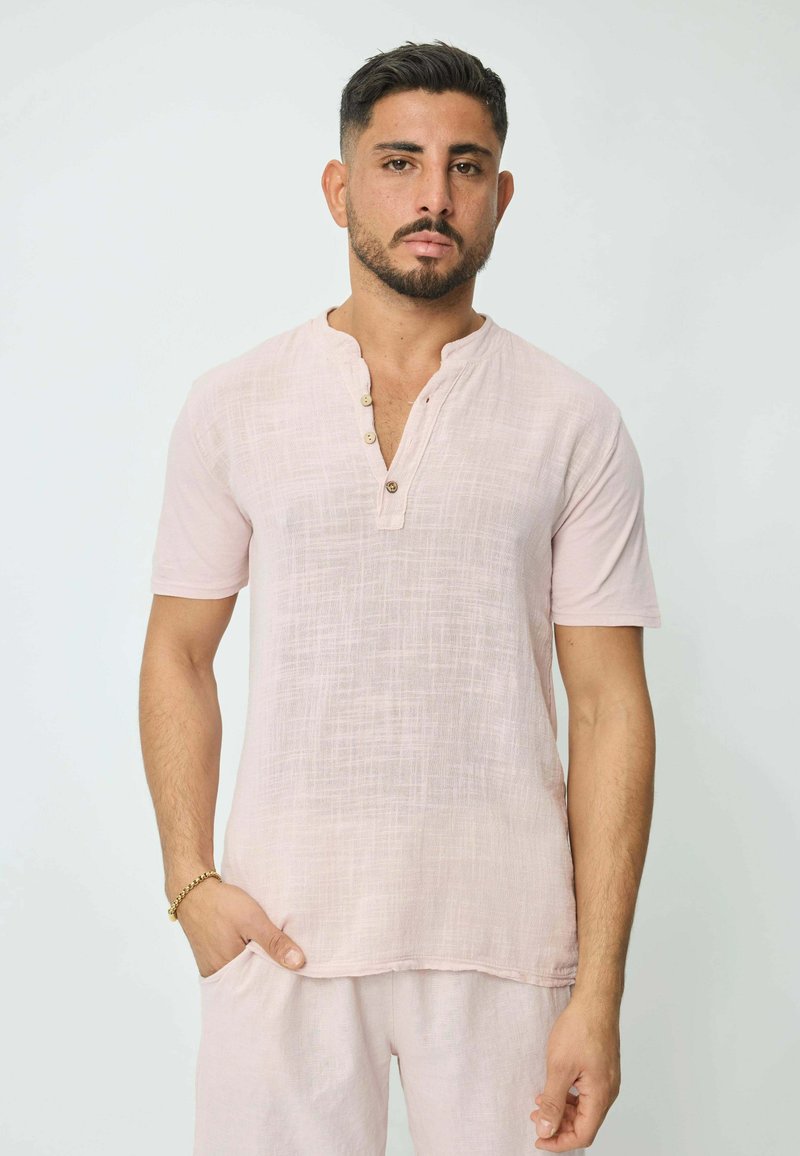 Chemise à manches courtes en lin rose clair avec une patte de boutonnage partielle et une texture subtile, portée avec un pantalon assorti.