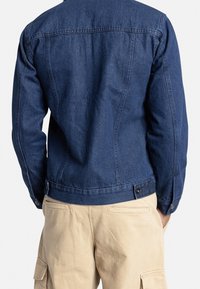 Homme portant une veste en denim bleu et un pantalon cargo beige, se tenant avec les mains dans les poches, dos à l'appareil photo sur fond blanc.