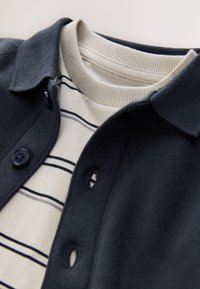 Tengerészkék gombos kardigán krémszínű csíkos hosszú ujjú ing fölött, bordázott mandzsettákkal és puha anyagokkal.