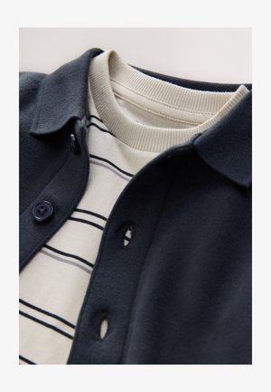 Marineblauw vest met knopen, gedragen over een crème gestreept shirt met lange mouwen, voorzien van geribbelde manchetten en zachte texturen.