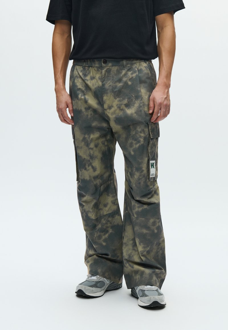 Pantalon cargo à motif tie-dye avec des nuances de vert et de marron, doté de poches latérales et d'un patch logo distinct sur la cuisse.