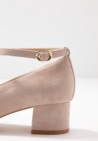 Scarpa beige in suede con tacco a blocco e cinturino alla caviglia. Dettaglio della fibbia dorata, superficie liscia, punta arrotondata e tacco corto e robusto.
