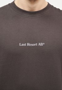 Άτομο που φοράει σκούρο γκρι μπλουζάκι με λευκό κείμενο "Last Resort AB®" στο κέντρο του στήθους.