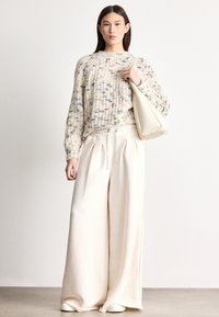 Pulover gros tricotat, crem cu pete albastre, asortat cu pantaloni largi crem și o geantă bej. Textură moale, croială lejeră, design minimalist.