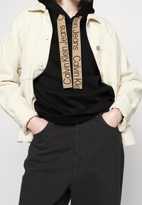 Jasnobeżowa kurtka dżinsowa na czarnej bluzie z kapturem z brązowymi sznurkami, z napisem "Calvin Klein Jeans", w połączeniu z ciemnymi spodniami.