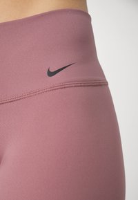 Leggings violets taille haute en tissu texturé et extensible avec un logo Nike noir sur la ceinture. Une couture horizontale traverse le haut.