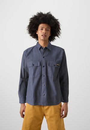 Camicia blu navy con bottoni, maniche lunghe, due taschini sul petto e un piccolo logo giallo; indossata con pantaloni corti gialli. Texture liscia, design casual.