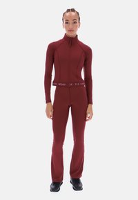 Vīnkrāsas sporta komplekts, kas sastāv no zipa augšdaļas ar garām piedurknēm un izplestām biksēm, izgatavots no elastīga, gluda materiāla ar zīmolu jostu.