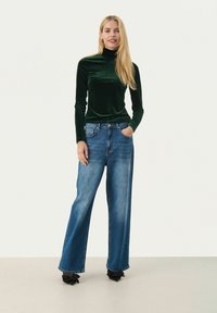 Grön sammetsmockneck-top i kombination med vida blå denimjeans. Accentuerad med en klassisk femficksdesign och svarta skor prydda med rosett.