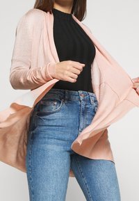 Ljusrosa cardigan med öppen front, ribbade ärmslut och nederkant. Bärs över en svart ribbad topp, tillsammans med blå denimjeans.