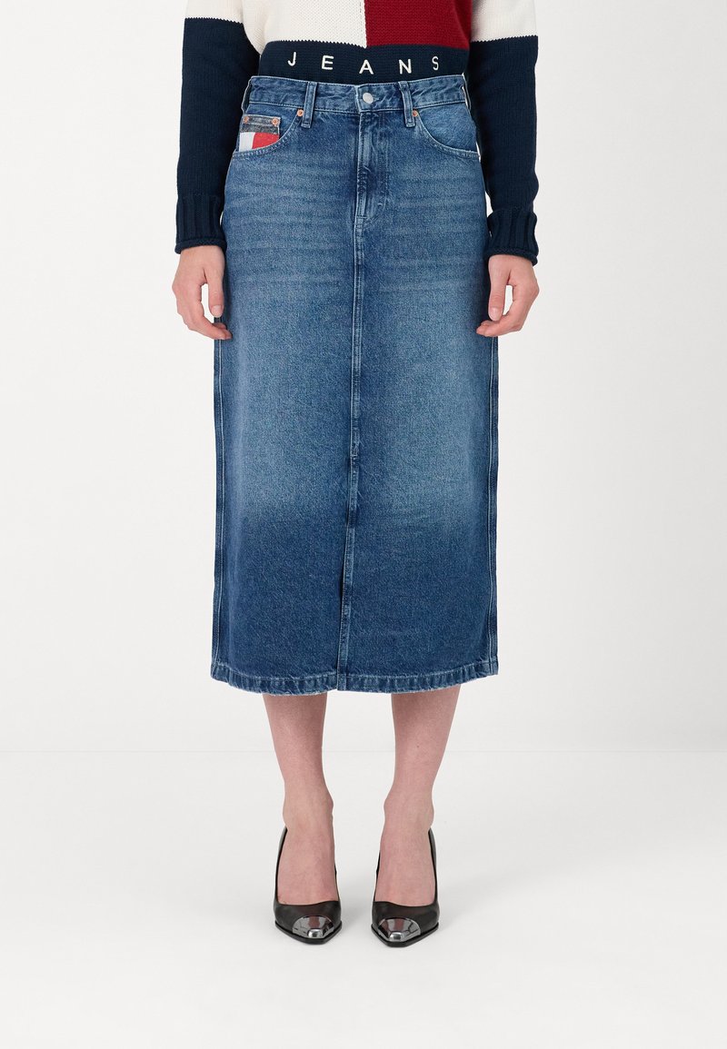 Denim midi svārki gaiši līdz vidēji zilā tonī ar priekšējo spraugu, piešūtu apakšmalu un kontrastējošiem kabatu akcentiem, saskaņoti ar melniem papēžiem.