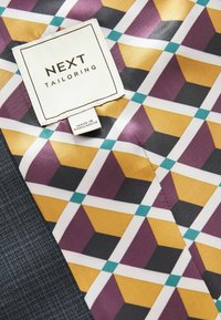 Étiquette de couture suivante sur un tissu à motifs présentant des formes géométriques en noir, jaune, violet et turquoise, à côté d'un matériau texturé bleu marine.