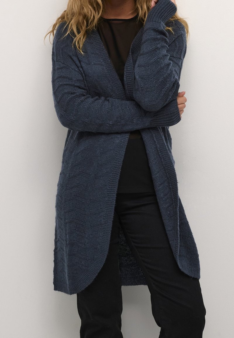 Cardigan lavorato a maglia di un blu scuro con un motivo testurizzato, con apertura sul davanti, maniche lunghe e una vestibilità comoda, indossato sopra un top e pantaloni neri.