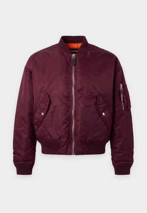 Homme portant une veste bomber bordeaux, un débardeur blanc, un jean bleu, des bottes noires, une casquette noire avec un logo orange, et une ceinture avec une boucle en forme de feuille d'érable rouge.