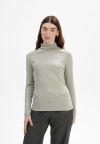 Langärmliges, hellgrünes Turtleneck-Oberteil aus weichem, glattem Stoff; taillierter Schnitt mit hohem Kragen und ärmellangen Ärmeln. Kombiniert mit dunklen Hosen.