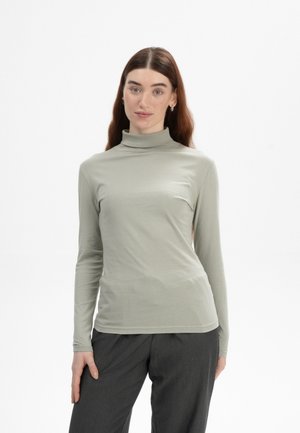 Langärmliges, hellgrünes Turtleneck-Oberteil aus weichem, glattem Stoff; taillierter Schnitt mit hohem Kragen und ärmellangen Ärmeln. Kombiniert mit dunklen Hosen.