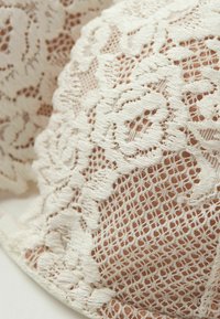 Spetsbralette med intrikata blommönster i krämfärg mot en naken bakgrund, med texturerade detaljer och mjuka kanter.