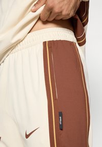 Κρέμ αθλητικές βερμούδες με κ�αφέ πλαϊνές πάνελ και κίτρινες ρίγες, ελαστική μέση και μια μικρή μαύρη υφαντή ετικέτα στο πλάι.