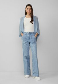 Lichtblauwe oversized cardigan over een witte aansluitende top, gecombineerd met lichtblauwe high-waisted jeans. Witte sneakers maken de outfit compleet.