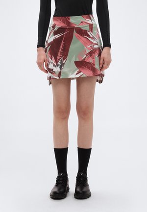 Kvinde iført en kort blomstret nederdel med røde og grønne blade, sort top med lange ærmer, sorte sokker og sorte snøresko, stående oprejst.