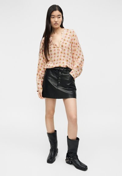 Γυναίκα με μακριά σκούρα μαλλιά που φοράει ροζ μπλούζα με λουλούδια, μαύρη μίνι φούστα από δέρμα με κουμπιά και μαύρες μπότες μέχρι τη μέση της γάμπας με αλυσίδα ως διακόσμηση.