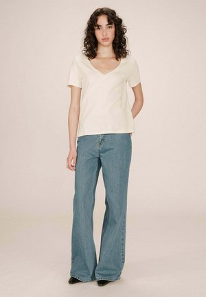 T-shirt beige en col en V fait de tissu doux, associé à un jean large bleu clair. La tenue est complétée par des chaussures noires.