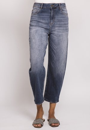 Frau trägt hoch taillierte, ausgewaschene blaue Jeans mit geradem Bein, kombiniert mit schwarz-weißen gewebten Plateau-Sandalen.