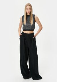 Pantaloni neri a gamba larga con pieghe abbinati a un top grigio attillato senza maniche con un maglione testurizzato. Il modello sta in piedi con le mani in tasca.