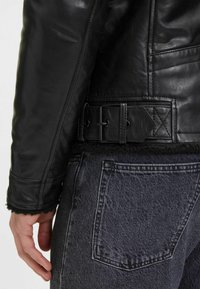 Giubbotto in pelle nera con finitura testurizzata, dotato di vita con cintura e morbido rivestimento in pile lungo i polsini. Jeans grigi scuri con tasca posteriore visibile.