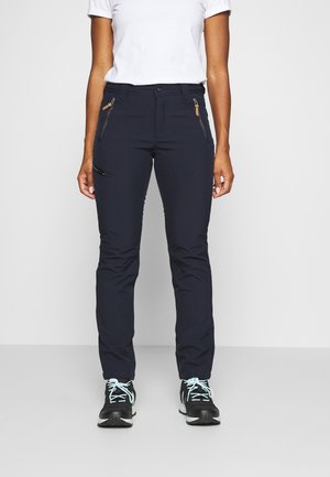 Personne portant un pantalon noir zippé, un t-shirt blanc et des baskets noires avec des lacets bleu clair, debout sur un fond uni.