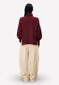 Person mit langen schwarzen Haaren, der einen lockeren weinroten Rollkragenpullover und weite beige Hosen trägt, steht mit dem Rücken zu einem weißen Hintergrund.