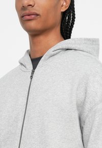 Közeli felvétel egy fonott hajú személyről, aki világosszürke, cipzáros kapucnis pulóvert visel egy sötét póló fölött, sima háttér előtt.