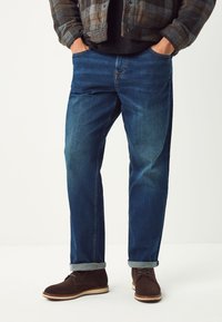 Dunkelblaue Jeans aus Denim mit lockerem Schnitt, leicht ausgeblichen, mit hochgekrempelten Säumen und Vordertaschen, kombiniert mit braunen Wildlederschuhen.