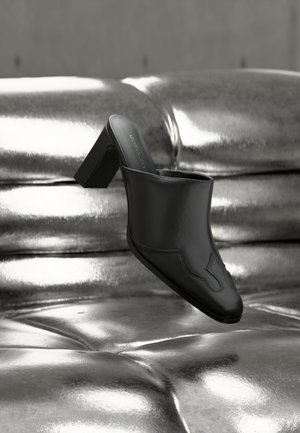 Mule en cuir noir avec talon bloc reposant sur une surface argentée brillante capitonnée, mettant en valeur son design élégant et ses détails incurvés.