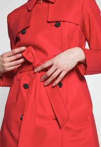 Gros plan sur des mains nouant la ceinture d'un trench-coat rouge vif avec des boutons noirs et des détails à rabat.