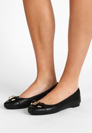 Sorte læder balletflats med sløjfe og guldbeslag båret på bare fødder mod en ensfarvet hvid baggrund.