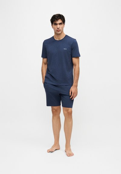 Homme debout, pieds nus, portant une chemise à manches courtes bleu marine et un short assorti sur un fond clair uni.