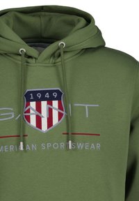 Πράσινο φούτερ με κουκούλα κατασκευασμένο από απαλό ύφασμα, με εμβληματική λεπτομέρεια στο μπροστινό μέρος που παρουσιάζει μια ασπίδα, κόκκινες και λευκές ρίγες, καθώς και κορδόνια διακόσμησης.