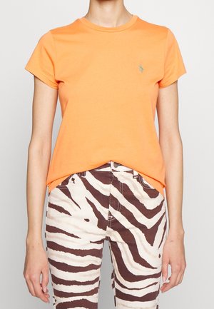 Person trägt ein kurzärmeliges oranges T-Shirt und hochtaillierte Hosen mit braunen und weißen Zebrasteifen, steht vor einem einfarbigen Hintergrund.