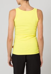 Gul randig linne med en åtsittande design, som har en rund halsringning och en liten röd etikett vid fållen, bärs tillsammans med svarta byxor.