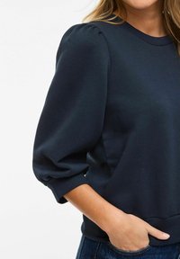 Femme portant un haut bleu marine à manches ballon trois-quarts, une main dans la poche d'un jean en denim, coupé aux épaules et à la taille.